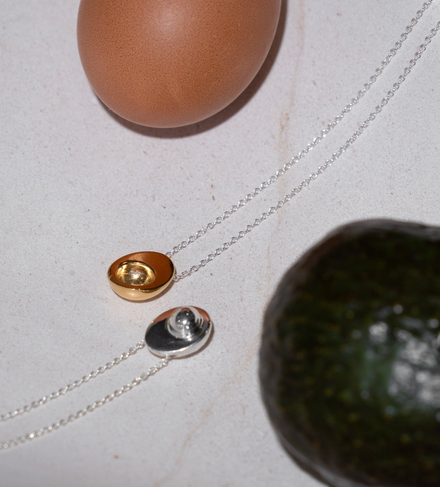 AGMES 0.925 sterling silver avocado and 18k gold vermeil egg necklace on white surface next to an egg and an avocado.