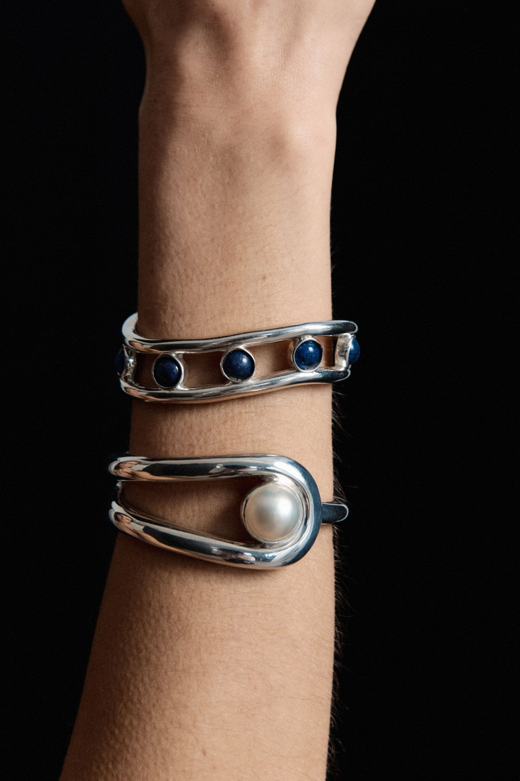Model wearing AGMES .925 sterling silver and lapis wavy sculptural Cara Cuff, and freshwater pearl rounded Becky Cuff. On black background.