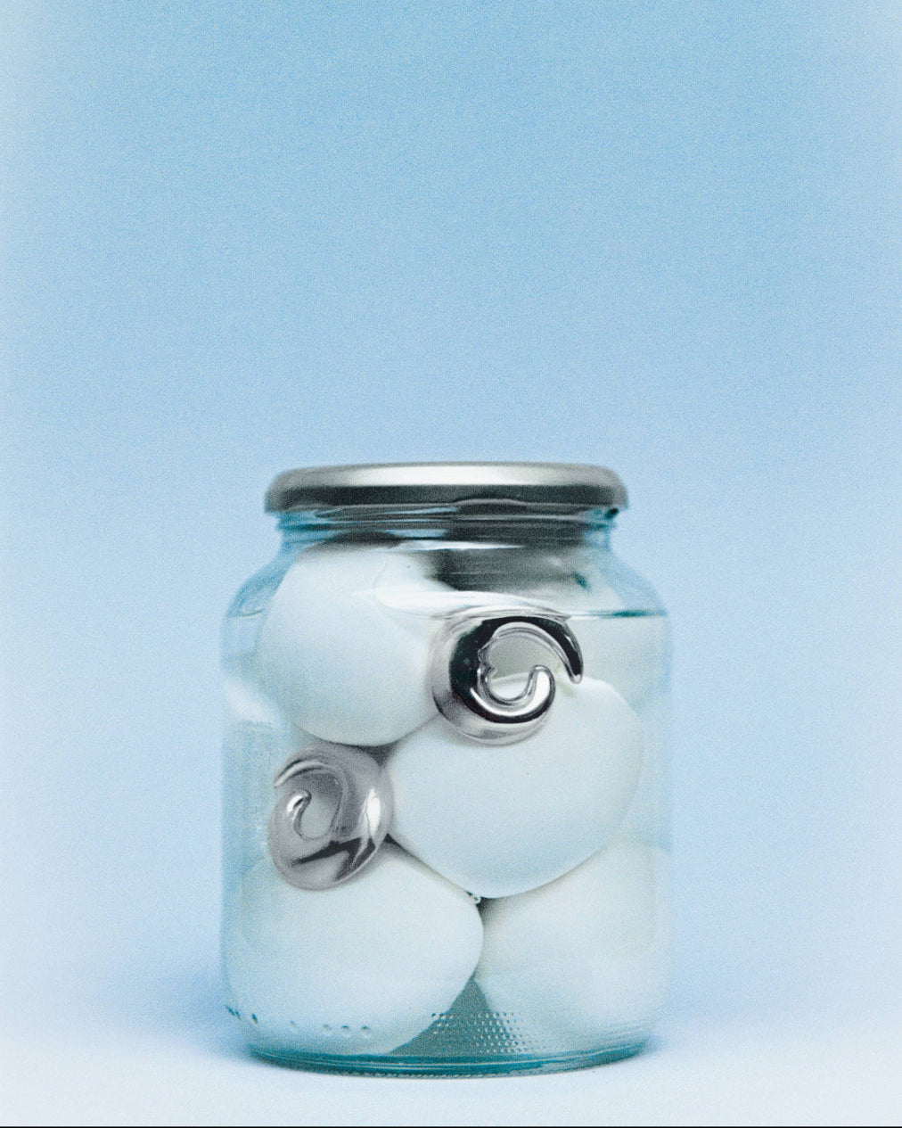 AGMES .925 sterling silver sculptural spiral heart round Nadia Earrings in glass jar with water and white eggs. On blue background.