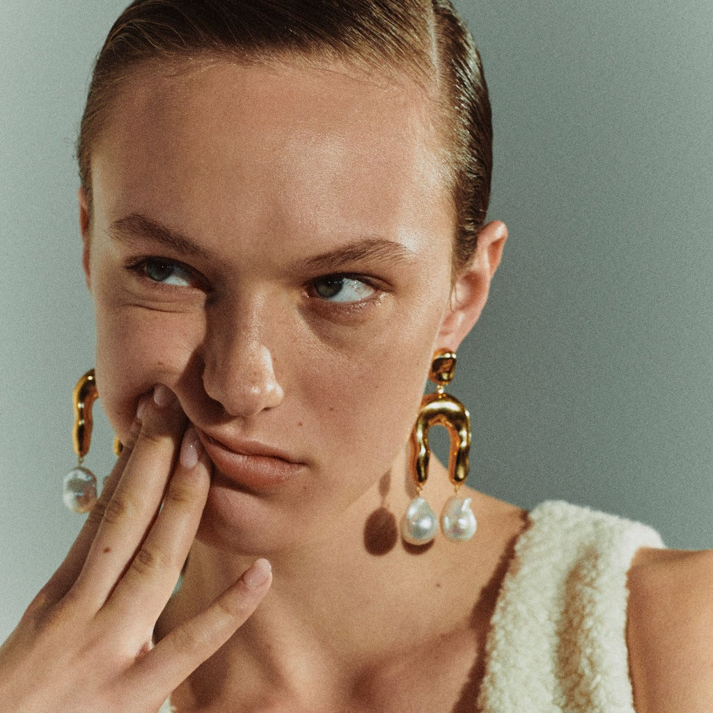 Woman wearing AGMES 18k gold vermeil earrings with sculpted wishbone and freshwater pearl drop.
