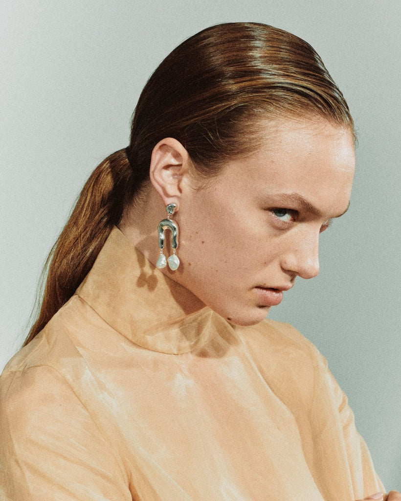 Woman wearing AGMES 0.925 sterling silver earrings with sculpted wishbone and freshwater pearl drop.