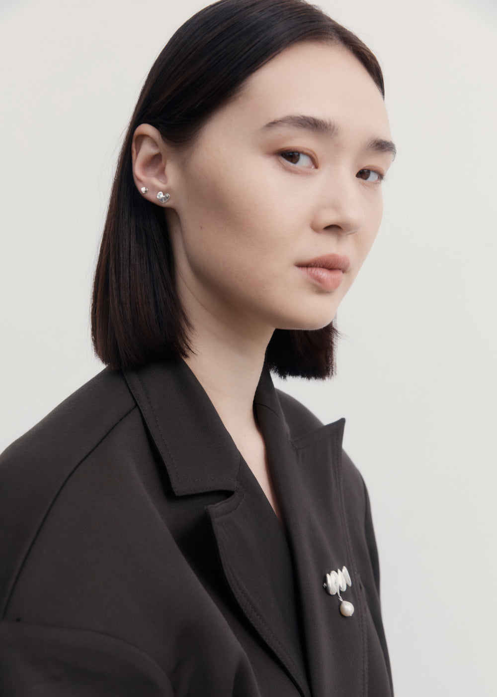 Woman wearing AGMES .925 Sterling Silver Joy Brooch with Baroque Pearl Drop on black blazer. Also wearing 0.925 Sterling Silver Mini Sculpted Heart Studs.