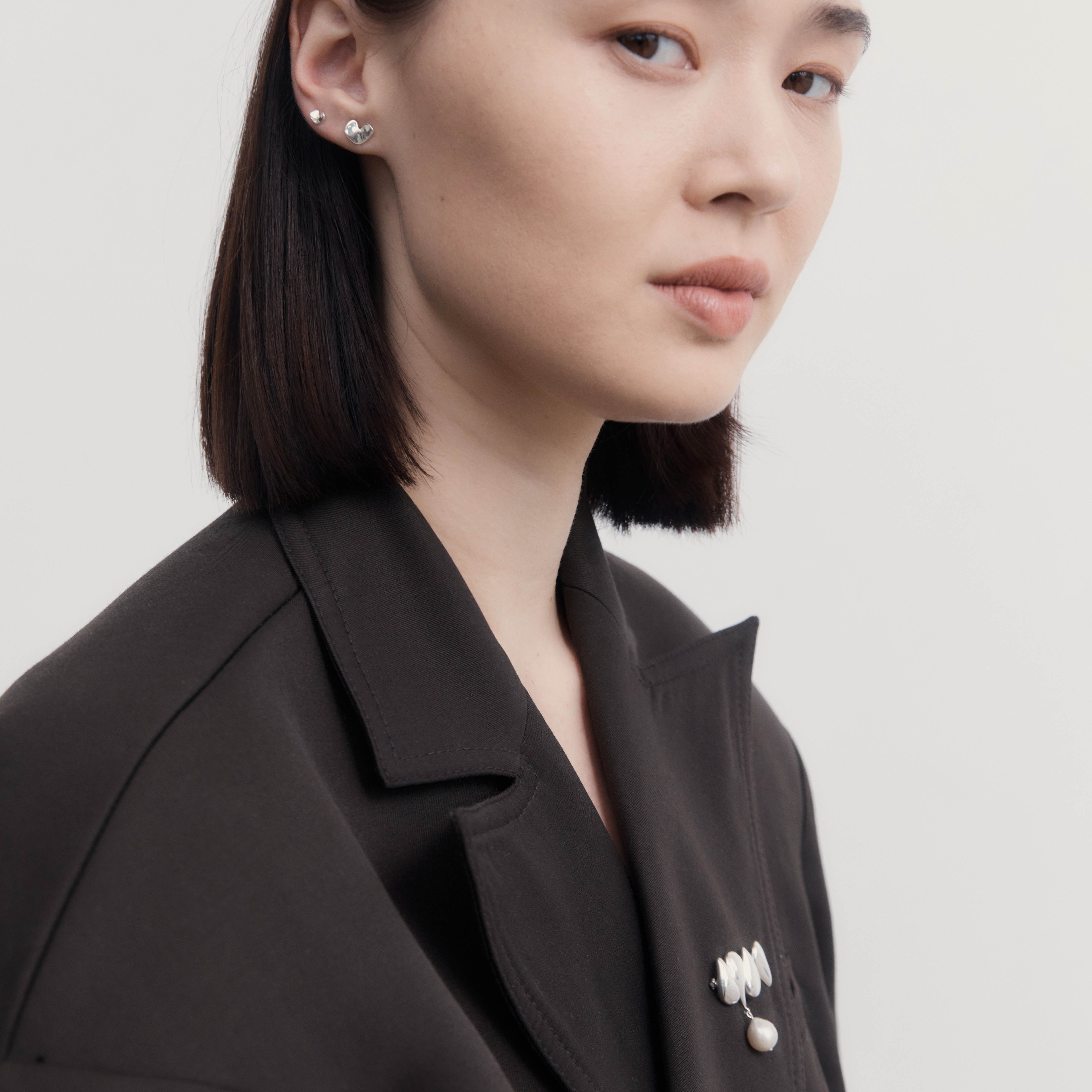 Woman wearing AGMES .925 Sterling Silver Joy Brooch with Baroque Pearl Drop on black blazer. Also wearing 0.925 Sterling Silver Mini Sculpted Heart Studs.
