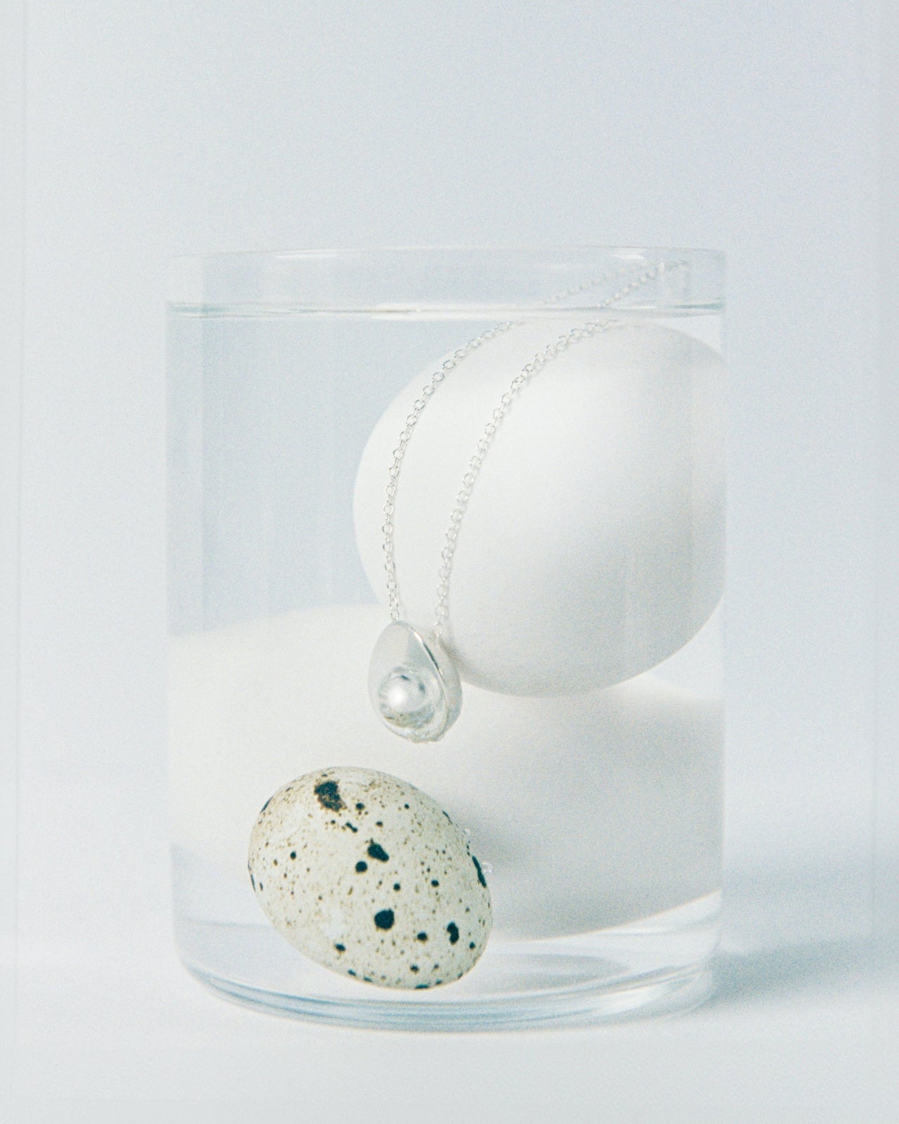 AGMES .925 sterling silver avocado pendant necklace in glass of water with white and speckled eggs. On white background.