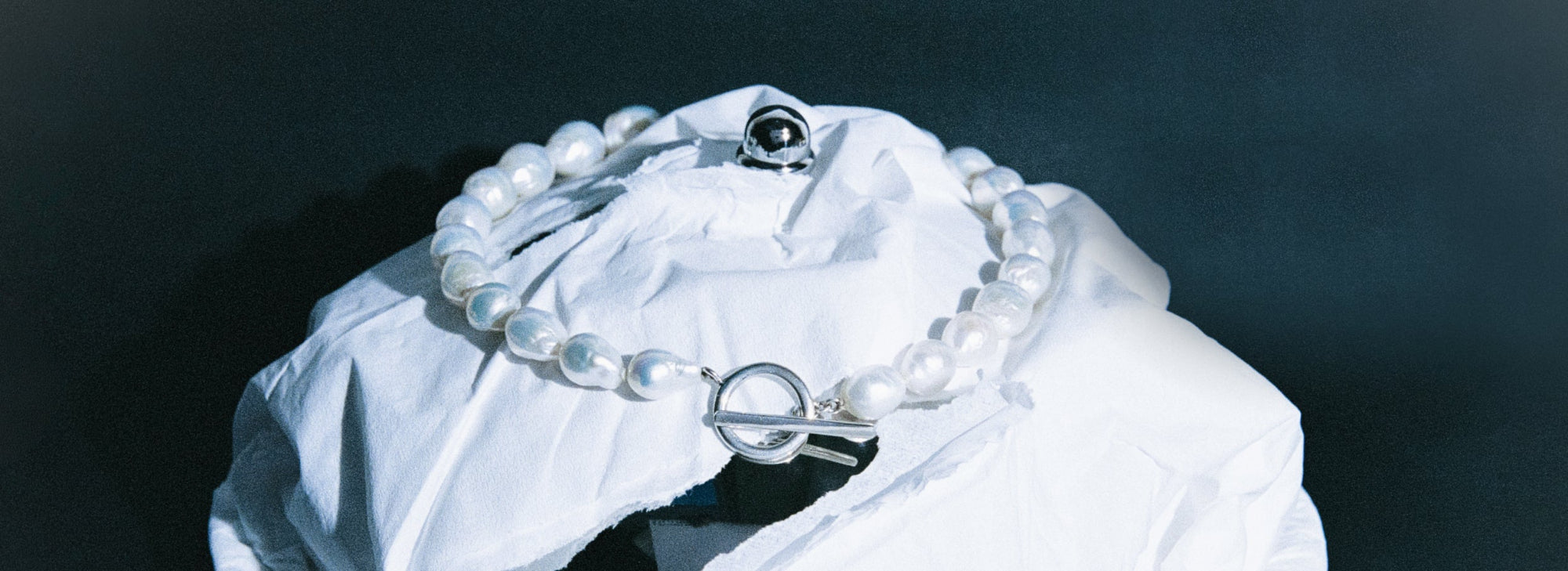 AGMES .925 sterling silver and freshwater pearl Baroque Pearl Necklace on white paper wrapped dish. Dark blue background.