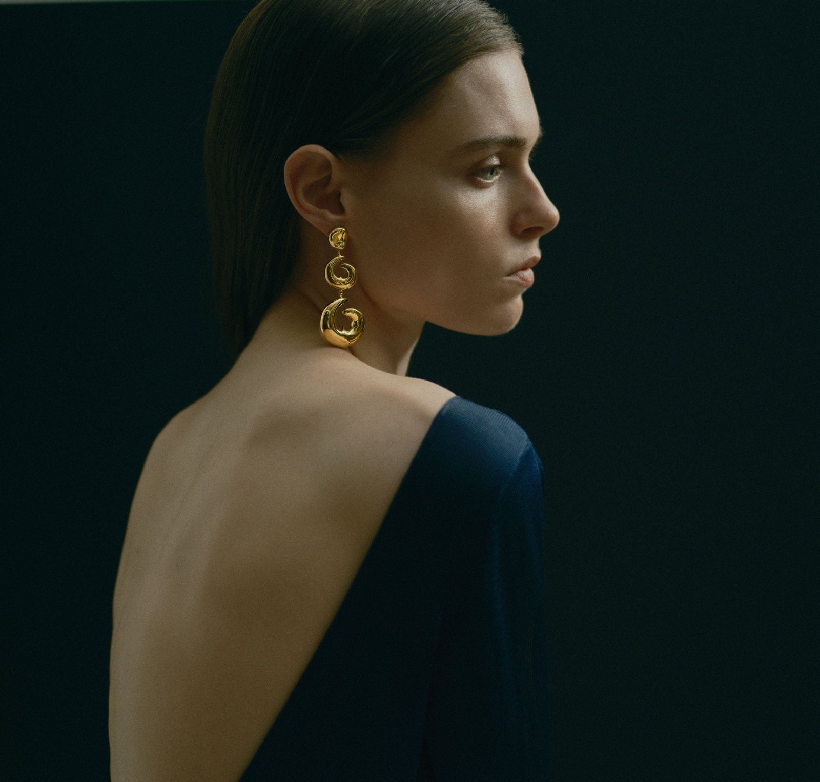 Model wearing AGMES 18k gold vermeil sculptural heart spiral Hilda Earrings. Black background.