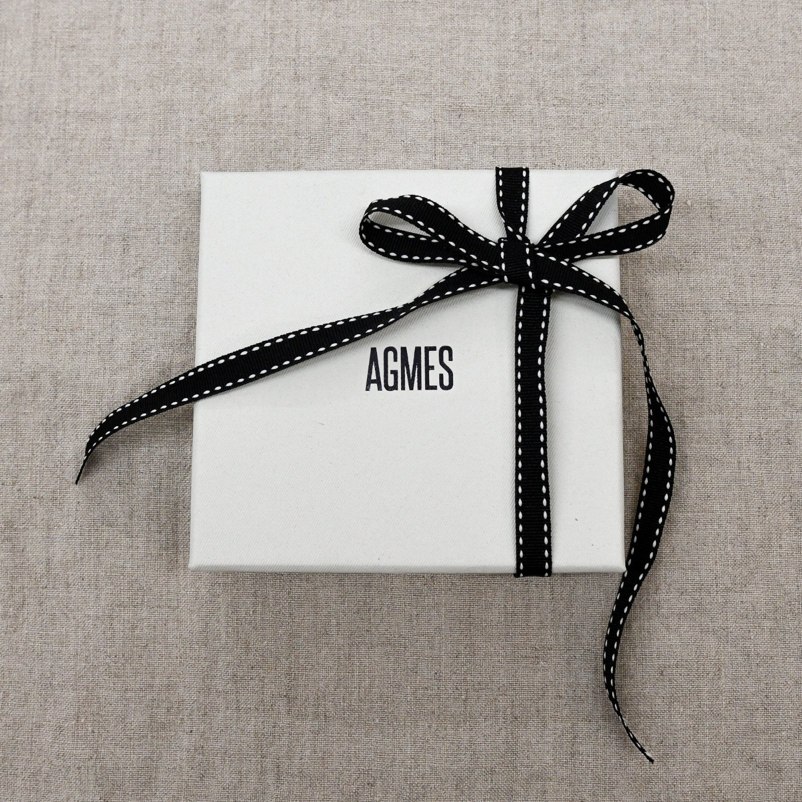 AGMES white logo gift box with black and white bow on tan linen background.