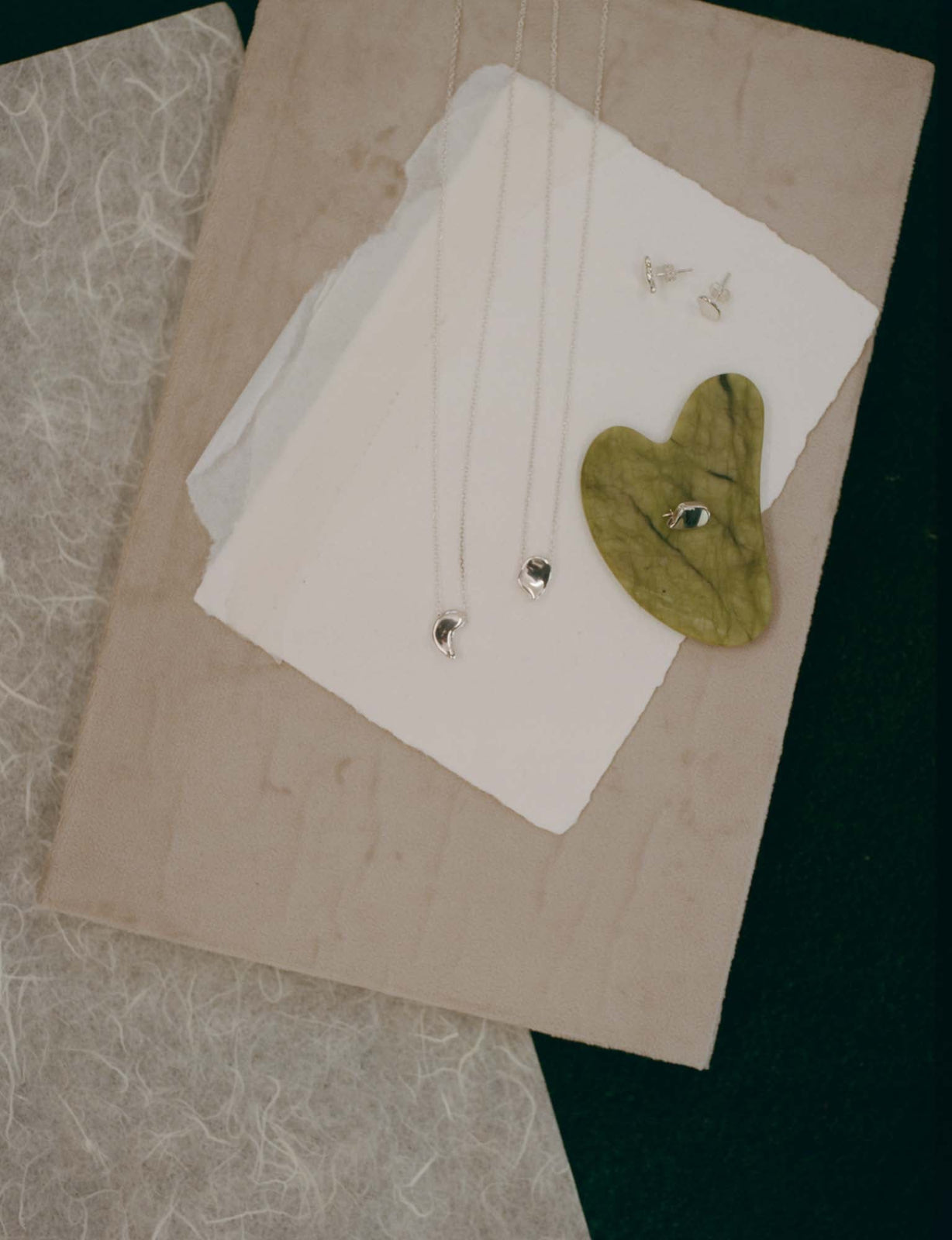 AGMES .925 sterling silver sculptural sum of parts heart pendant necklaces, mini brooch, and stud earrings. On green stone, white and green fabric background.