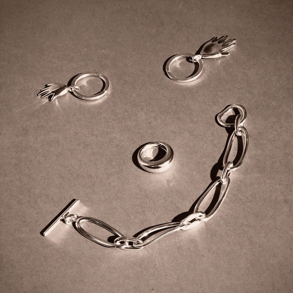 AGMES .925 sterling silver sculptural link Dion Bracelet, Domed Ring, and hand shaped Dali Keychains on grey background.